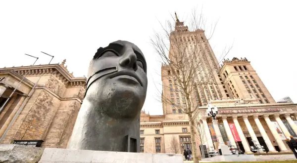 Monumentalna rzeźba głowy na tle Pałacu Kultury w Warszawie.